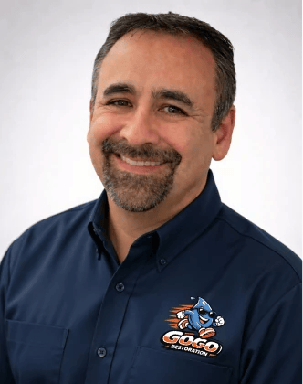 Smiling man with short dark hair and goatee wearing a navy blue "Go Go Restoration" shirt, posing against a plain light background—ready to tackle water mitigation and mold remediation projects.